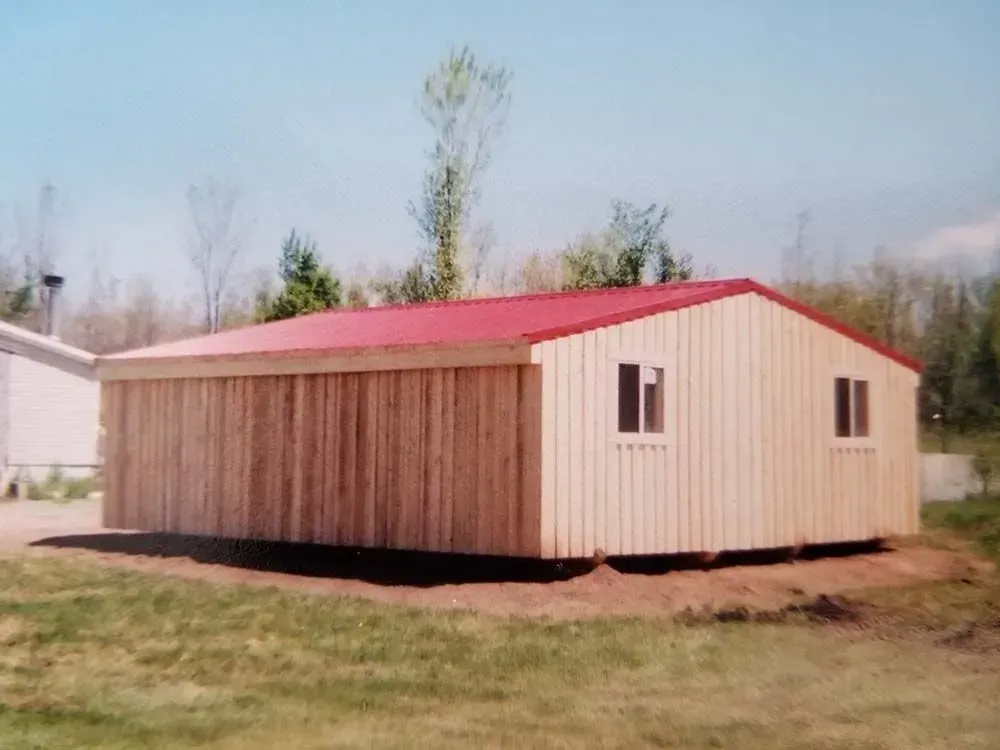 Wooden building with red roof, two windows, and light-colored siding. Set on grass with trees in the background.