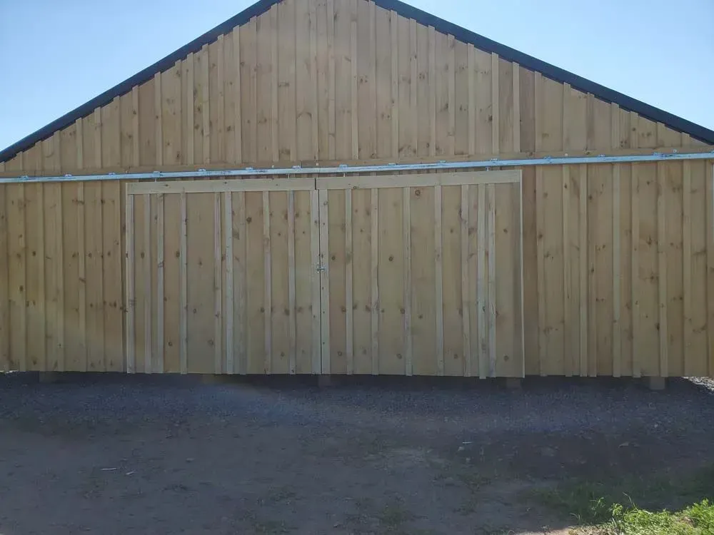 Wooden barn with sliding doors, a black roof, and gravel ground.
