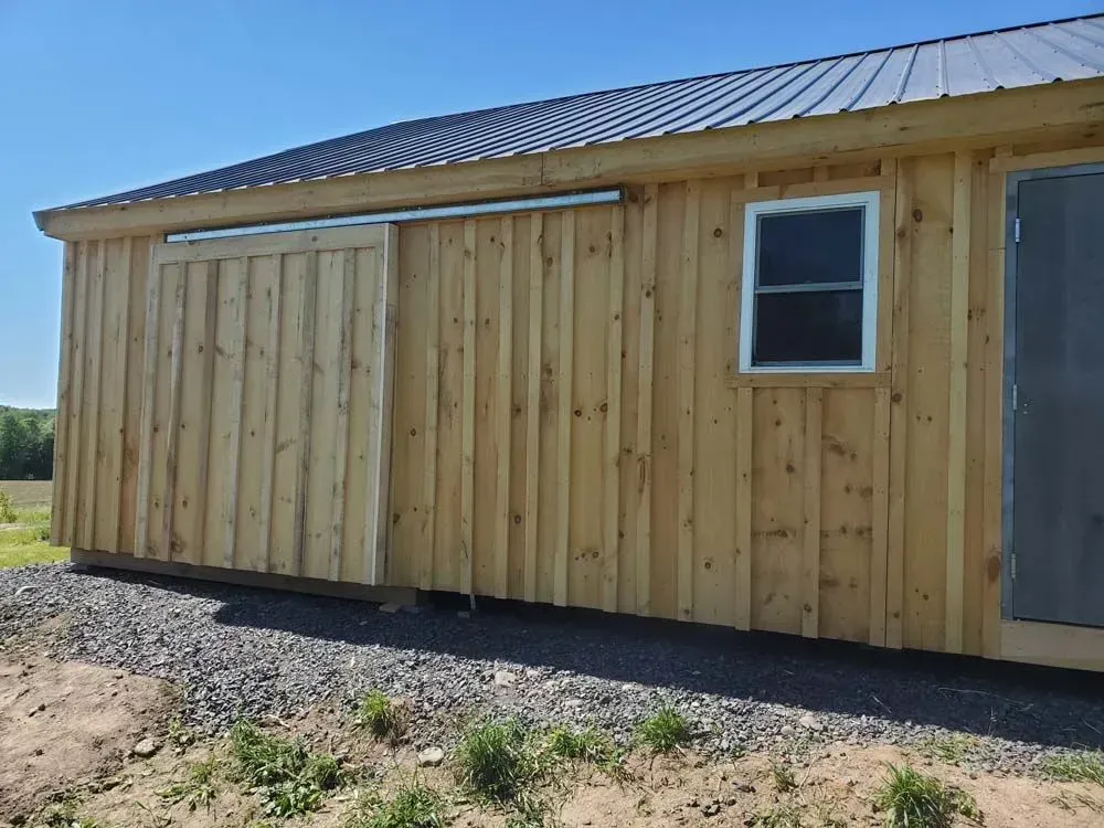 Wooden shed with a sliding door, window, and metal roof against a blue sky.