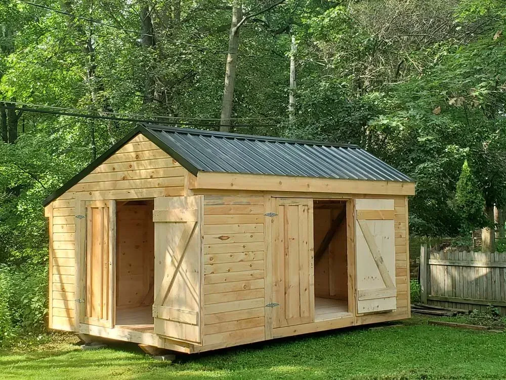 Wooden shed with corrugated metal roof, two doorways with shutters, set in a grassy yard.
