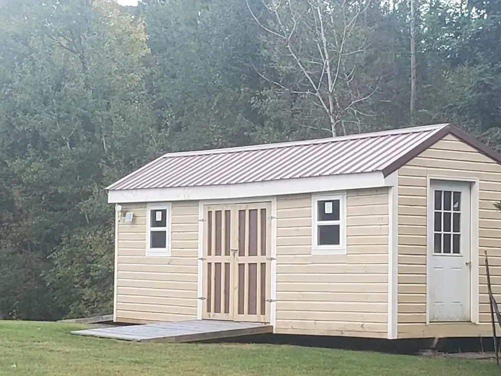 Tan shed with brown roof, white door, and windows; set in front of green trees.