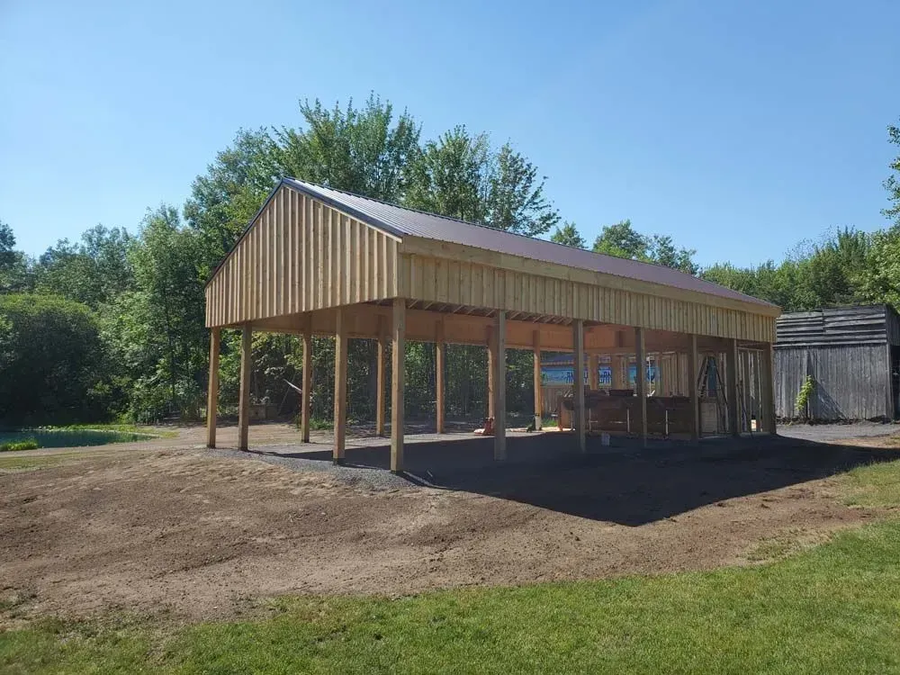Wooden open-air pavilion with a brown metal roof. Built on a gravel pad, surrounded by grass and trees.