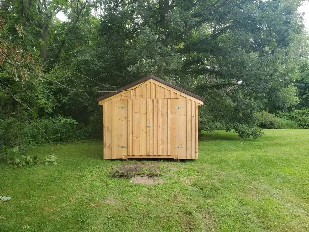 Wooden shed with brown roof on a green lawn, surrounded by trees.