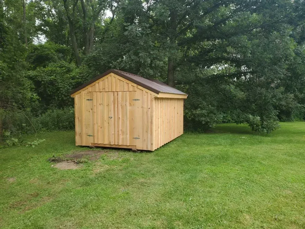 Wooden shed with a brown roof on a grassy lawn, surrounded by trees.