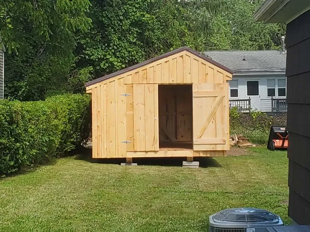 Wooden shed with open door on a green lawn, surrounded by trees and a house.