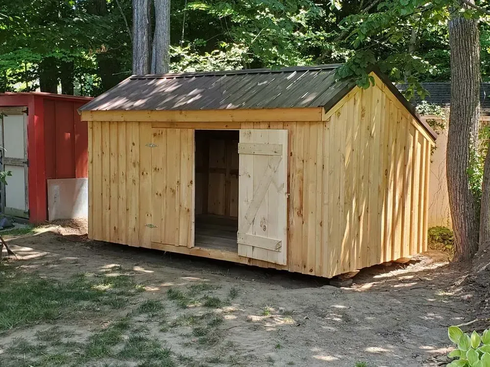 Wooden shed with open door, metal roof, and two wooden shutters. Located in a grassy yard with trees.