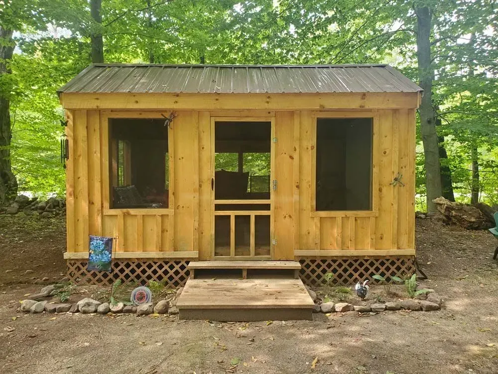 Small wooden cabin with screened door and windows, set in a wooded area.
