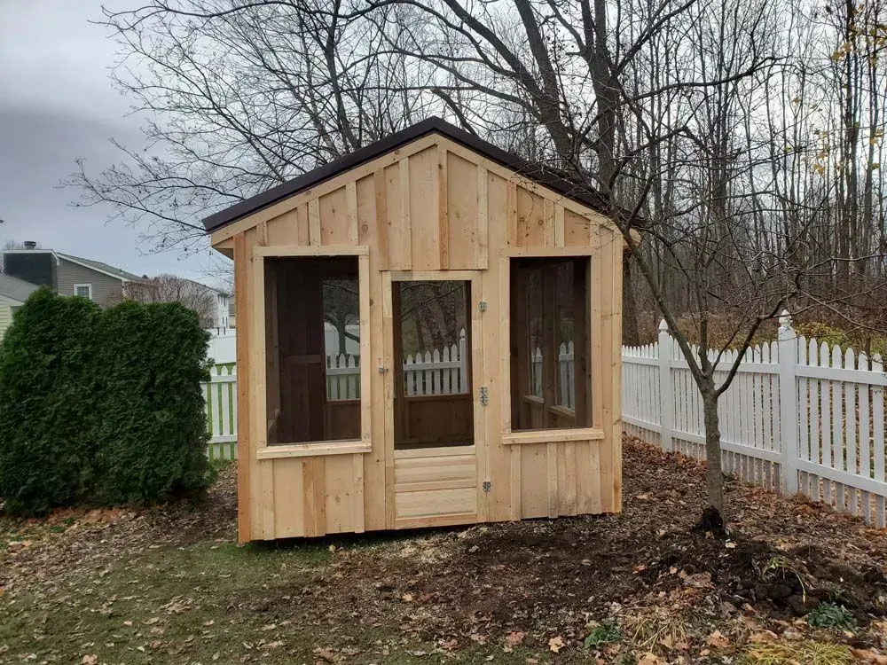 Wooden shed with screened windows and door in a backyard, brown roof, surrounded by trees and white fence.