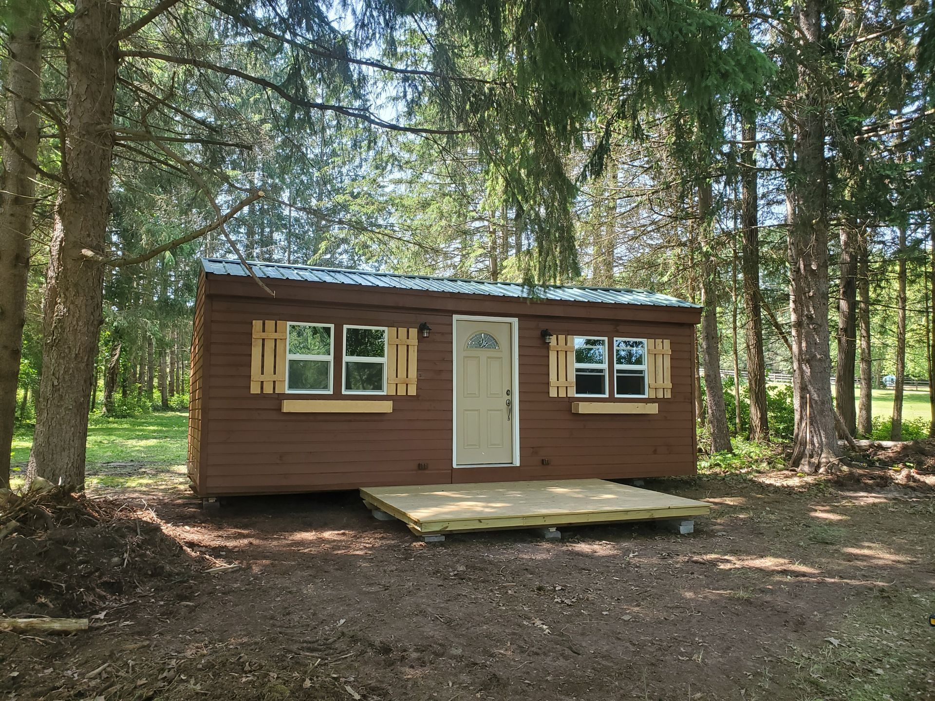 Wooden shed with open door, metal roof, and two wooden shutters. Located in a grassy yard with trees.