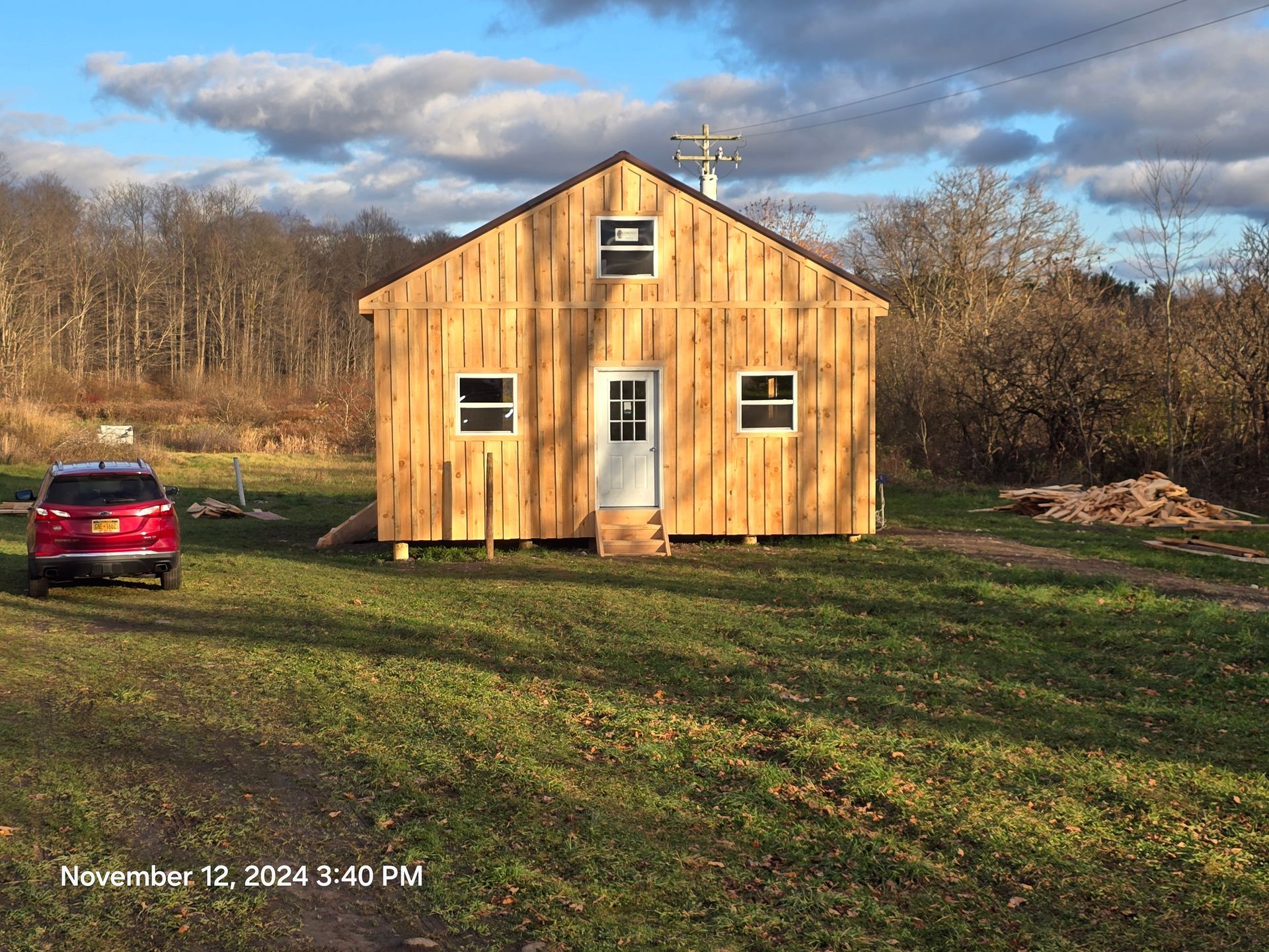 Tan shed with brown roof, white door, and windows; set in front of green trees.