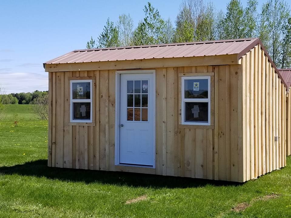 Wooden shed with white door and windows, brown roof, set in a grassy field on a sunny day.
