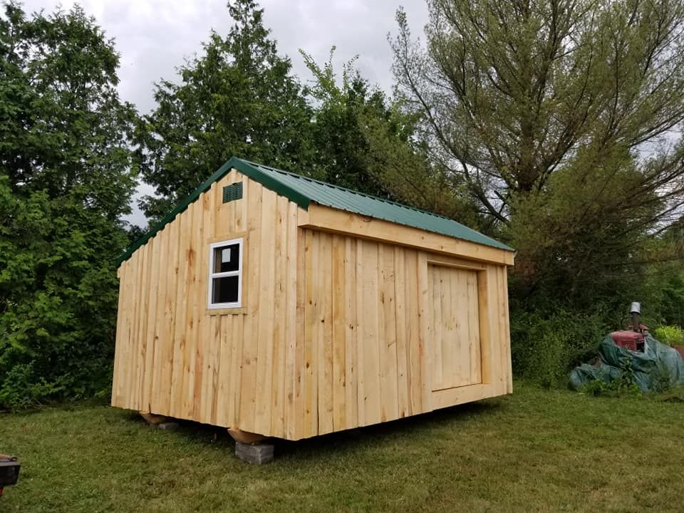 Wooden shed with green roof and a small window, sits on gray blocks in a grassy yard.
