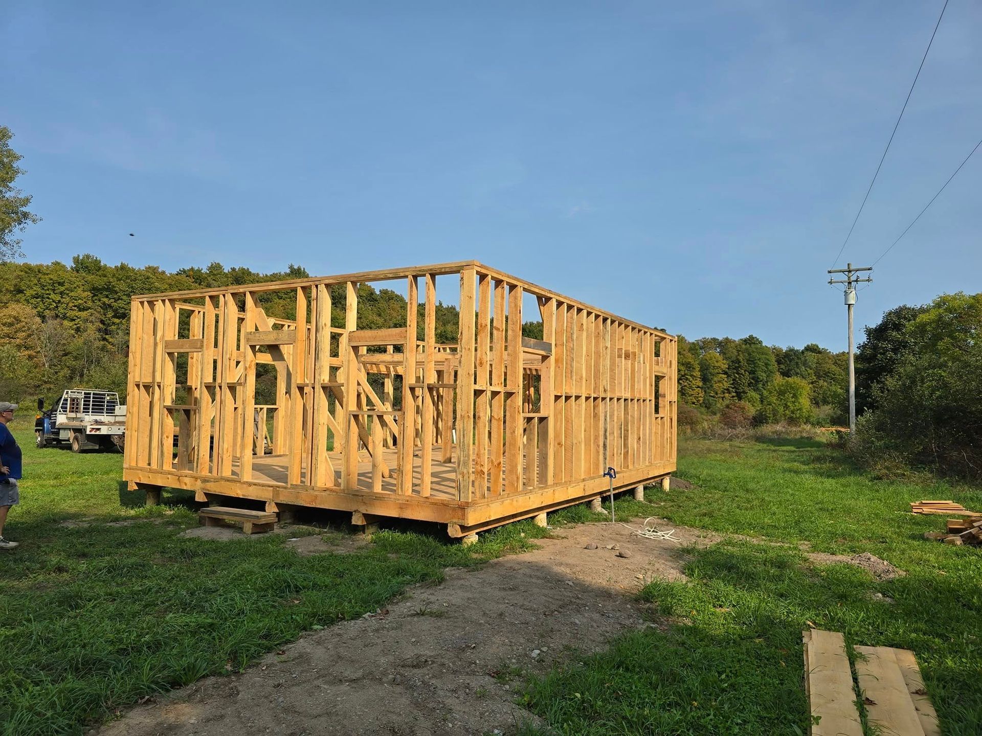 Wooden frame of a small building under construction on a grassy lot; trees in background.