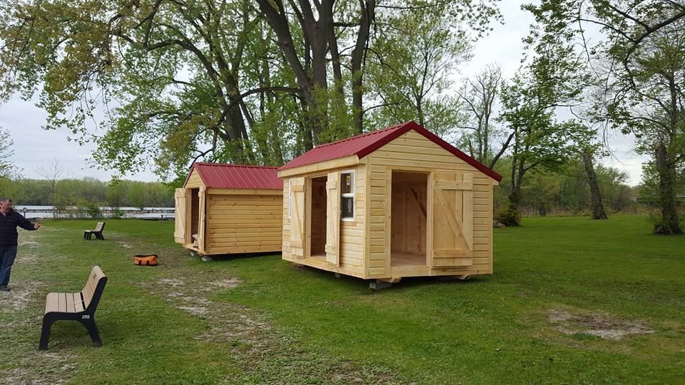 Two small wooden sheds with red roofs sit on grass. A person stands nearby, with trees in the background.