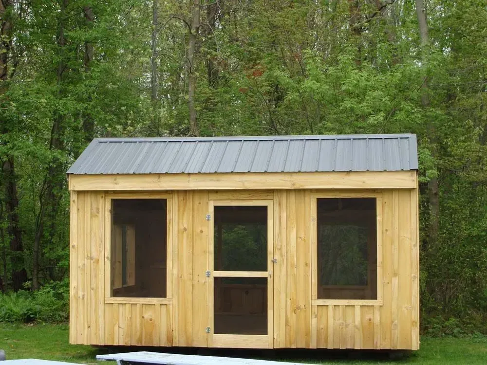Wooden shed with gray metal roof, three screened openings, set in a wooded area.