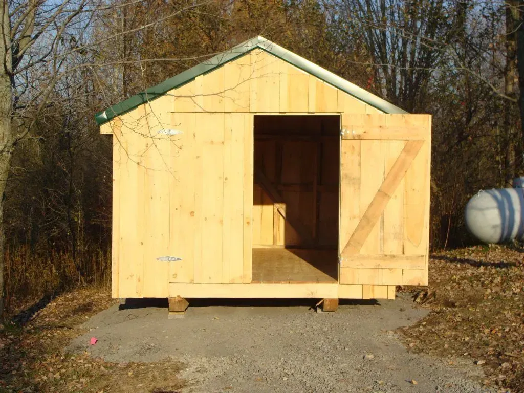Wooden shed with open door, green roof, sitting on blocks outdoors.