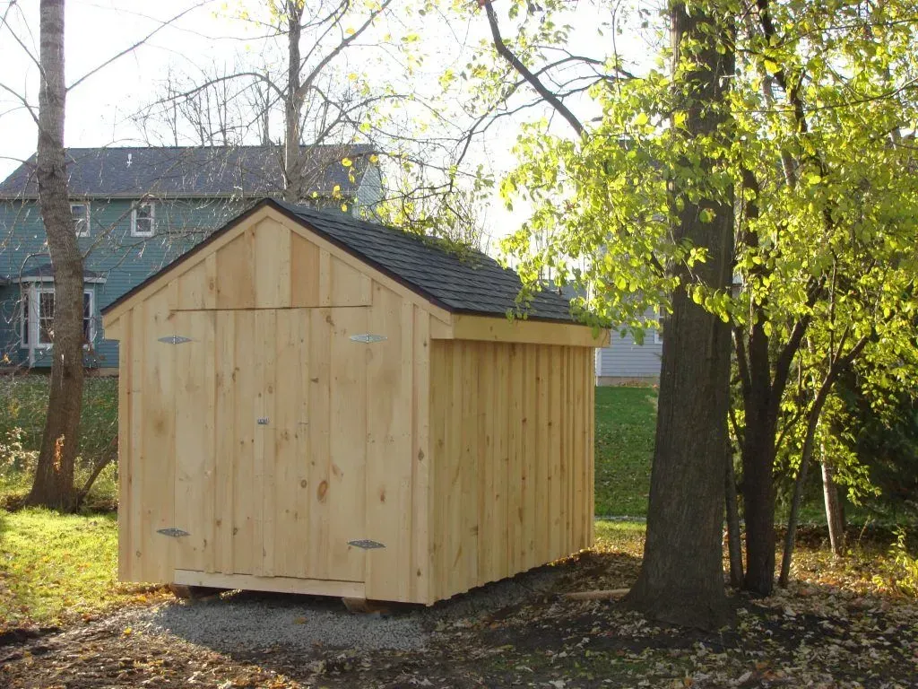 Wooden shed with black roof in a yard, trees in the background, sunny day.