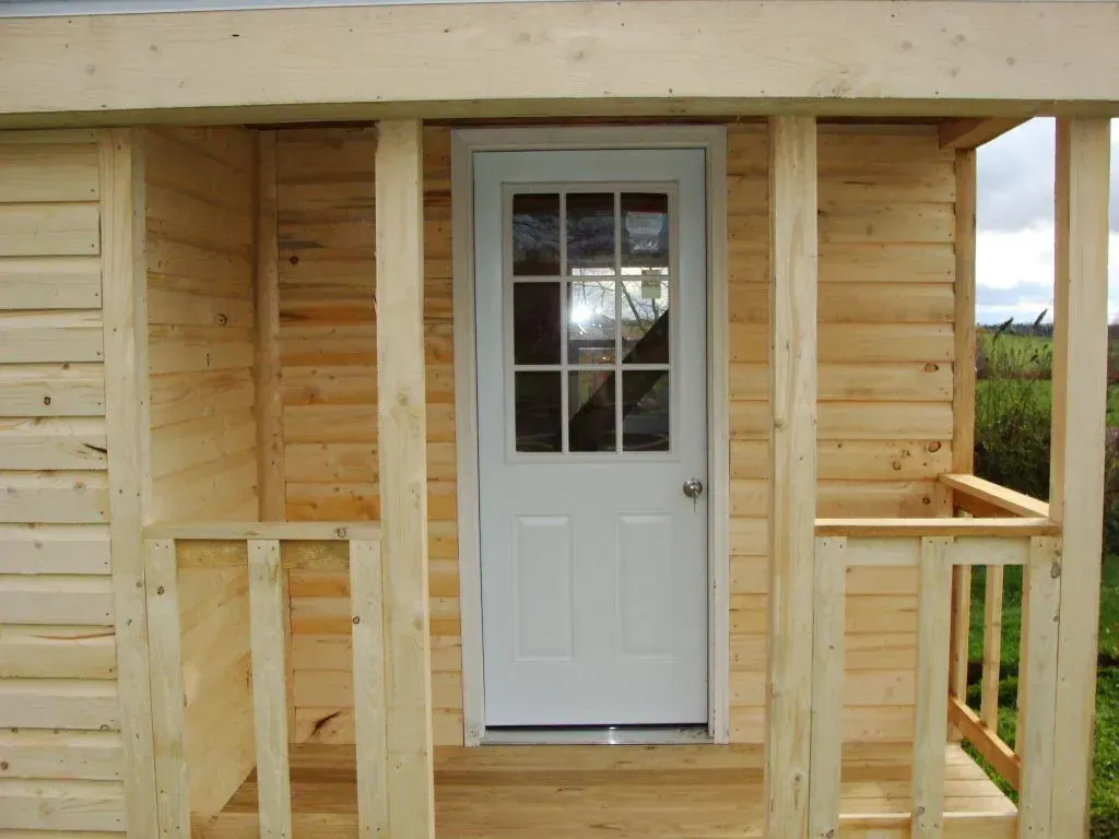 Wooden shed with white door and small porch; unfinished construction, outdoor setting.