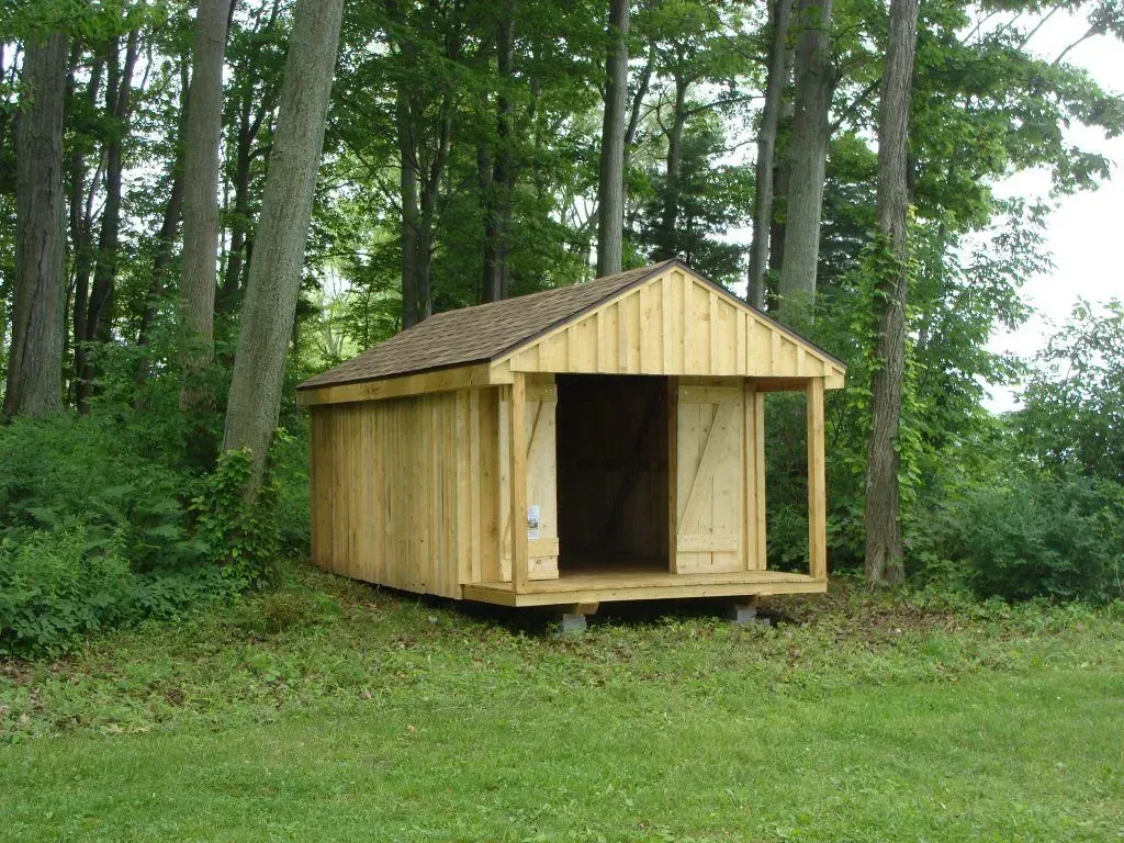 Wooden shed with an open doorway, set in a grassy area surrounded by trees.