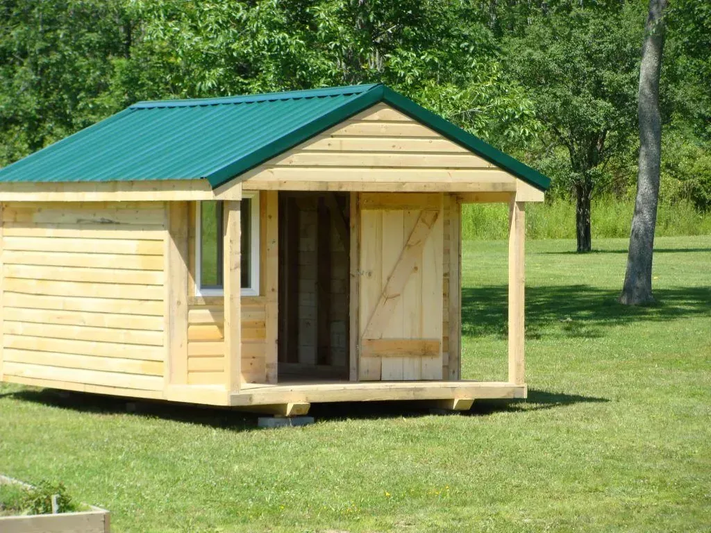 Small wooden cabin with green roof and porch in a grassy field, surrounded by trees.