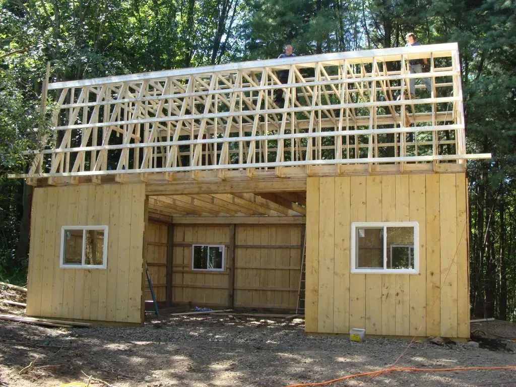 Barn under construction, with a wooden frame, windows, and unfinished roof. Forest background.