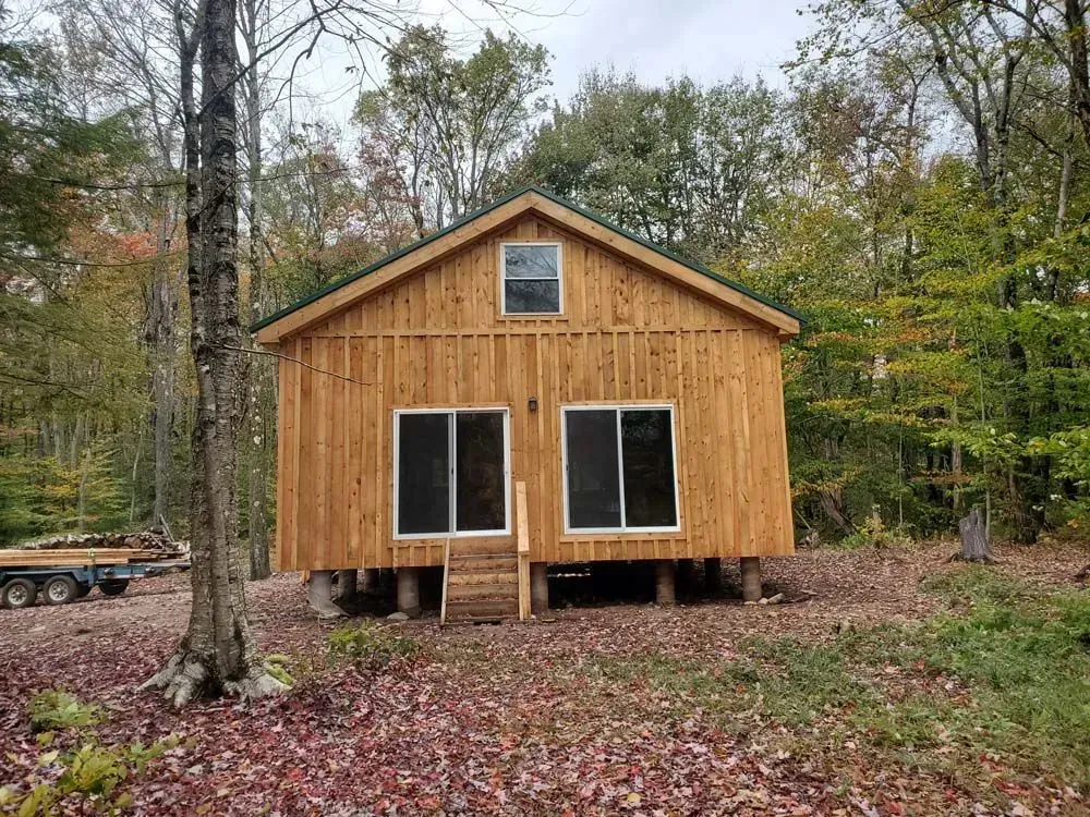 Small wooden cabin on stilts with sliding glass doors and a small window; set in a forest with fall foliage.