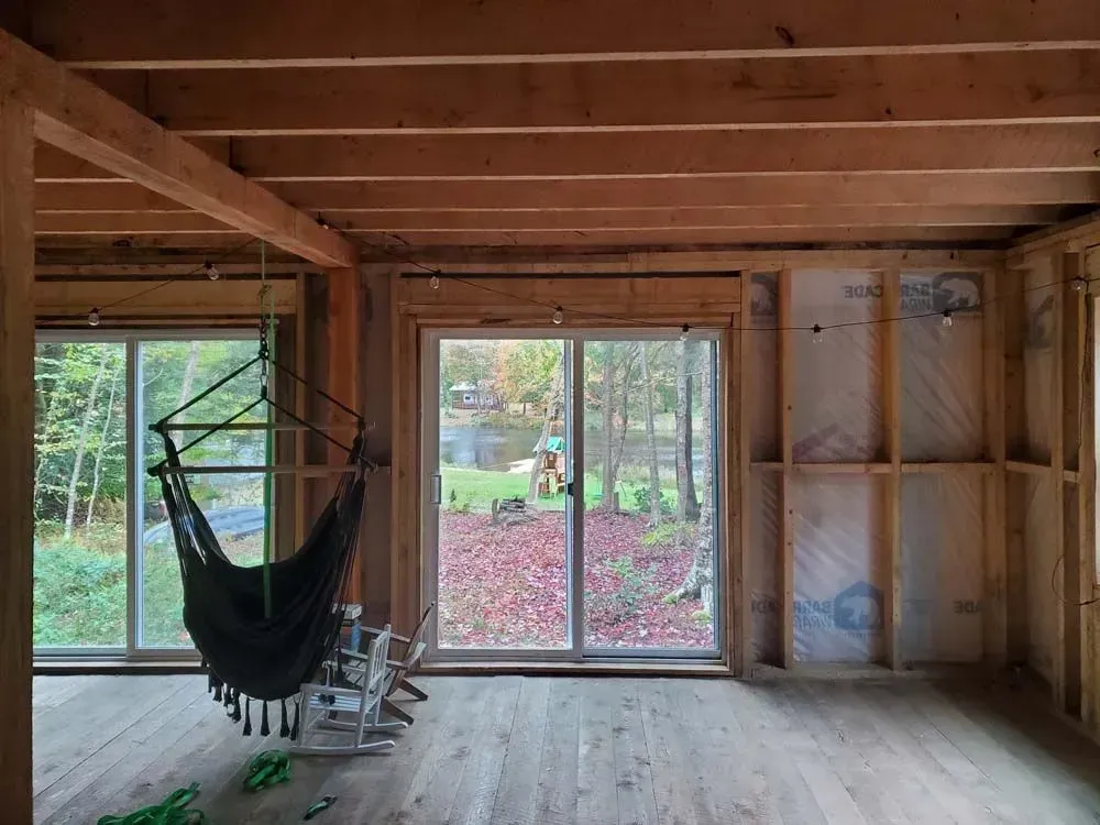 Interior view of a room under construction. Windows look out onto fall foliage. A hammock hangs inside.