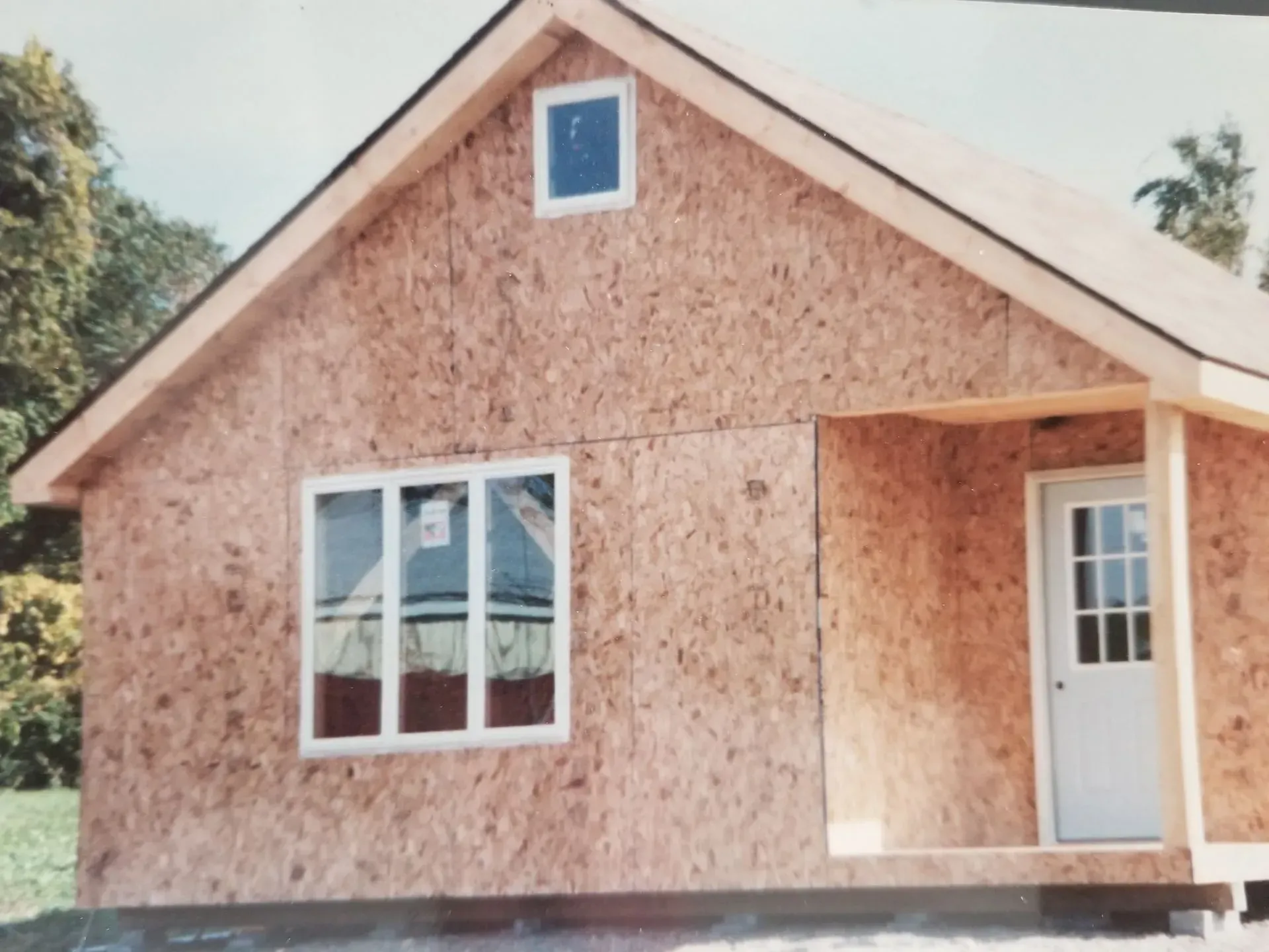 Unfinished cabin exterior with OSB siding, white trim, windows, and door, under construction in a grassy area.