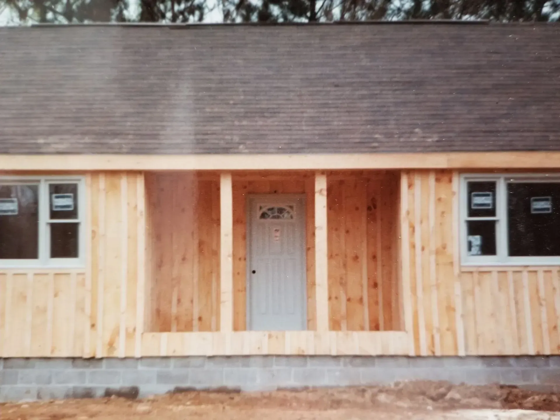 Unfinished house exterior with a front door, windows, and wooden siding.  Dark roof, light wood, grey foundation.