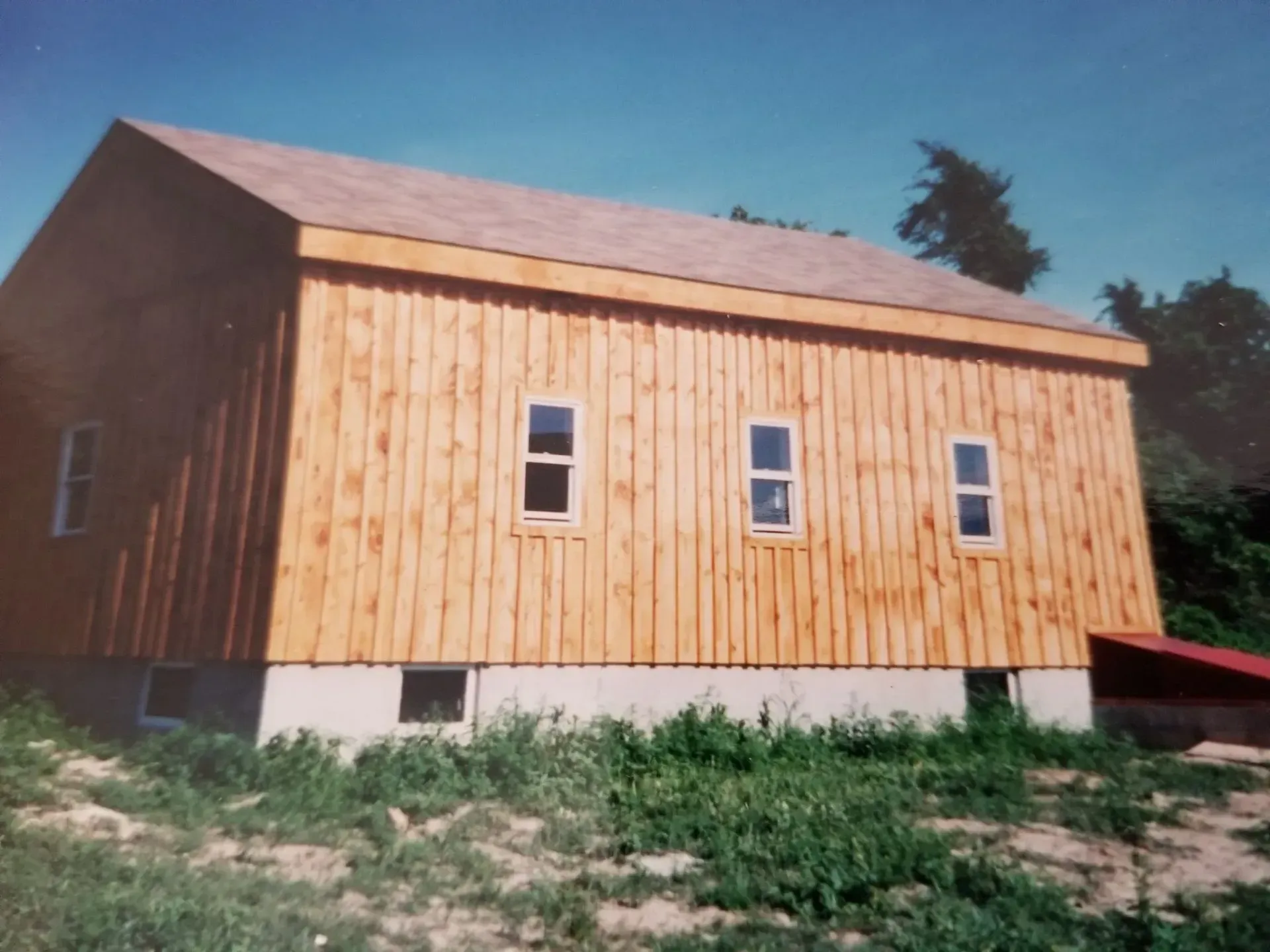 Wooden building with light-colored siding, small windows, and brown roof. Set in a field with green grass.