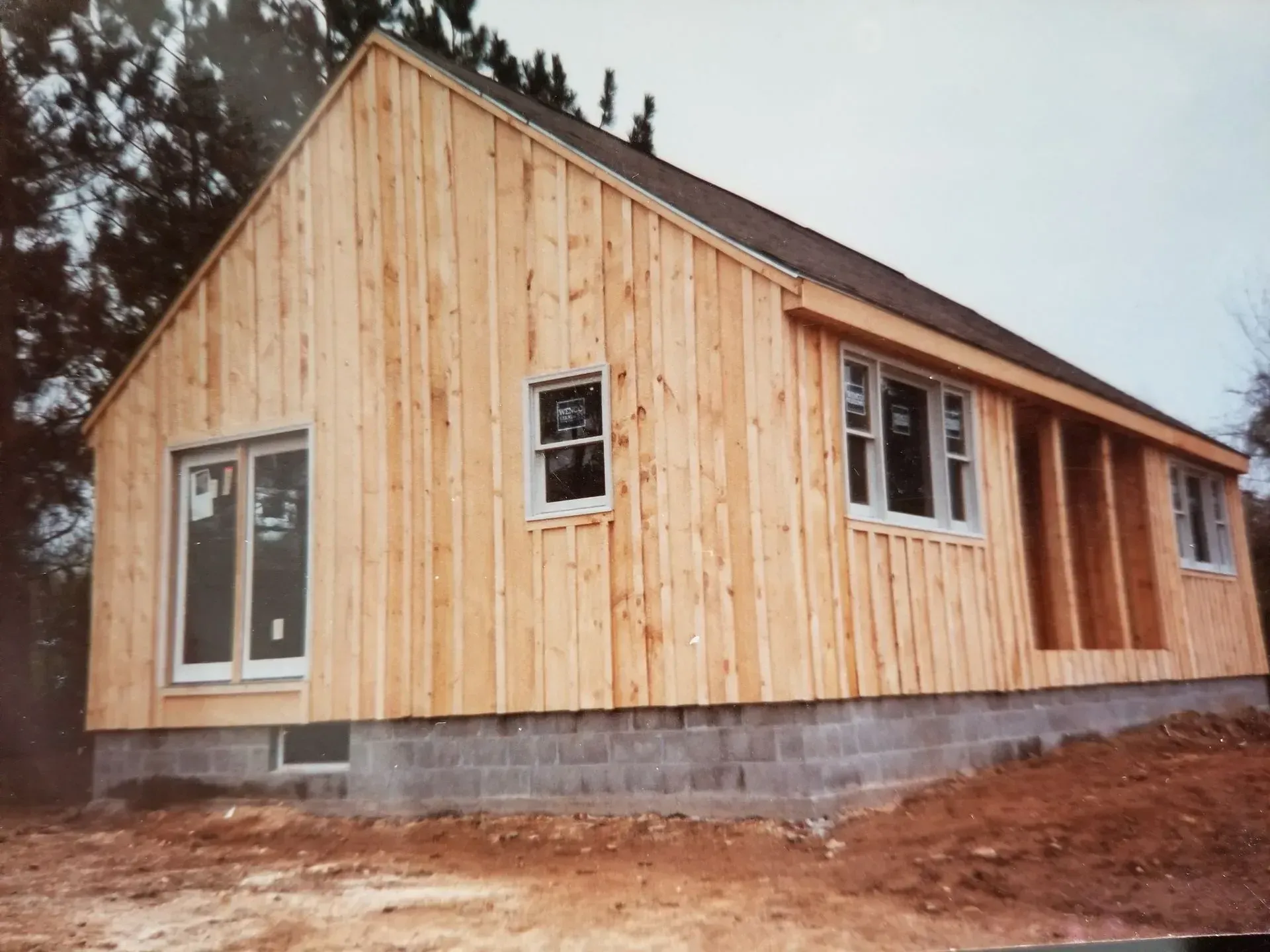 Unfinished wood-sided house on a cinder block foundation, windows installed, construction site outdoors.