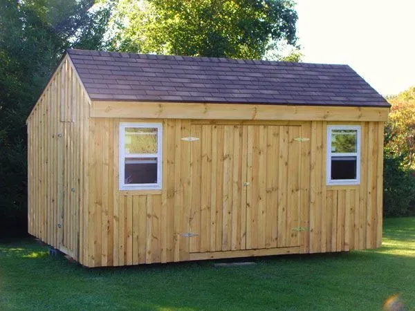 A wooden shed with a brown roof and white-framed windows sits on green grass in a yard.