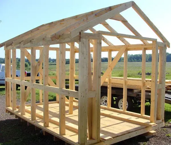 Wooden shed frame in progress outdoors on a trailer, with roof rafters and wall studs.