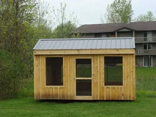Wooden shed with metal roof, three windows, and a screen door, set on a grassy lawn with a building in the background.