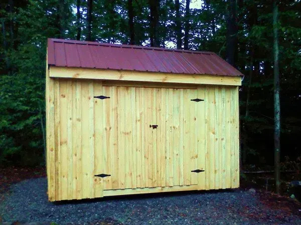 Wooden shed with a burgundy metal roof, in a wooded area.