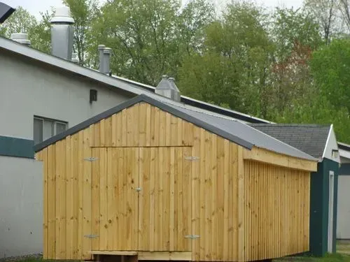 Wooden storage shed with a gray roof next to a building, set outdoors with trees in the background.