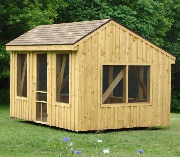 Wooden screened-in shed with a brown shingled roof sitting on green grass.