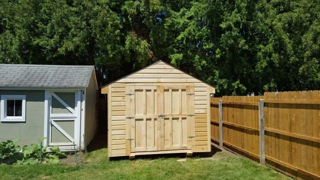 Two sheds side-by-side on grass. One is green, the other is wooden. A brown fence is to the right.