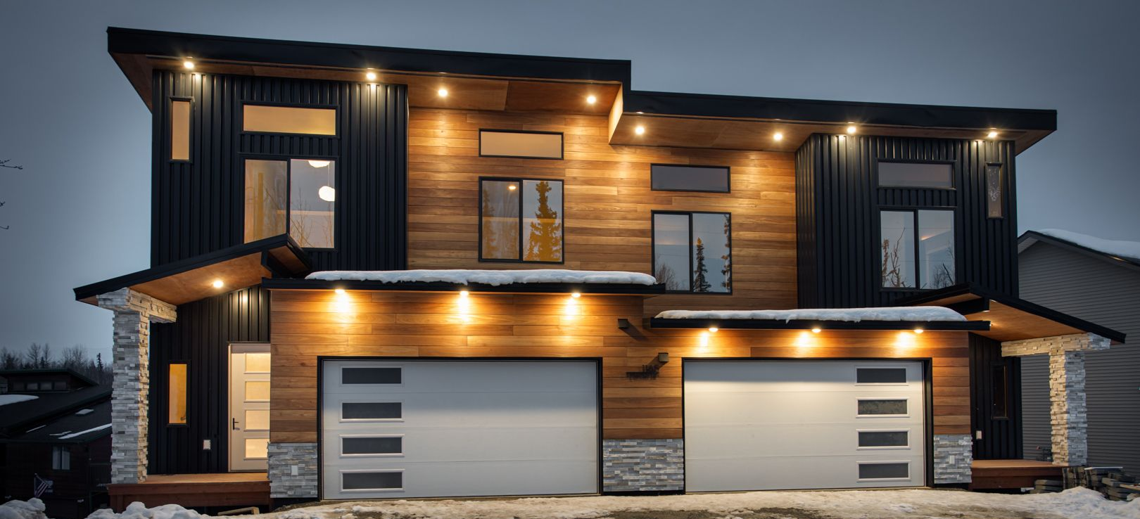 A modern two-story home with wood siding, black metal accents, and two garage doors at dusk, surrounded by light snow.