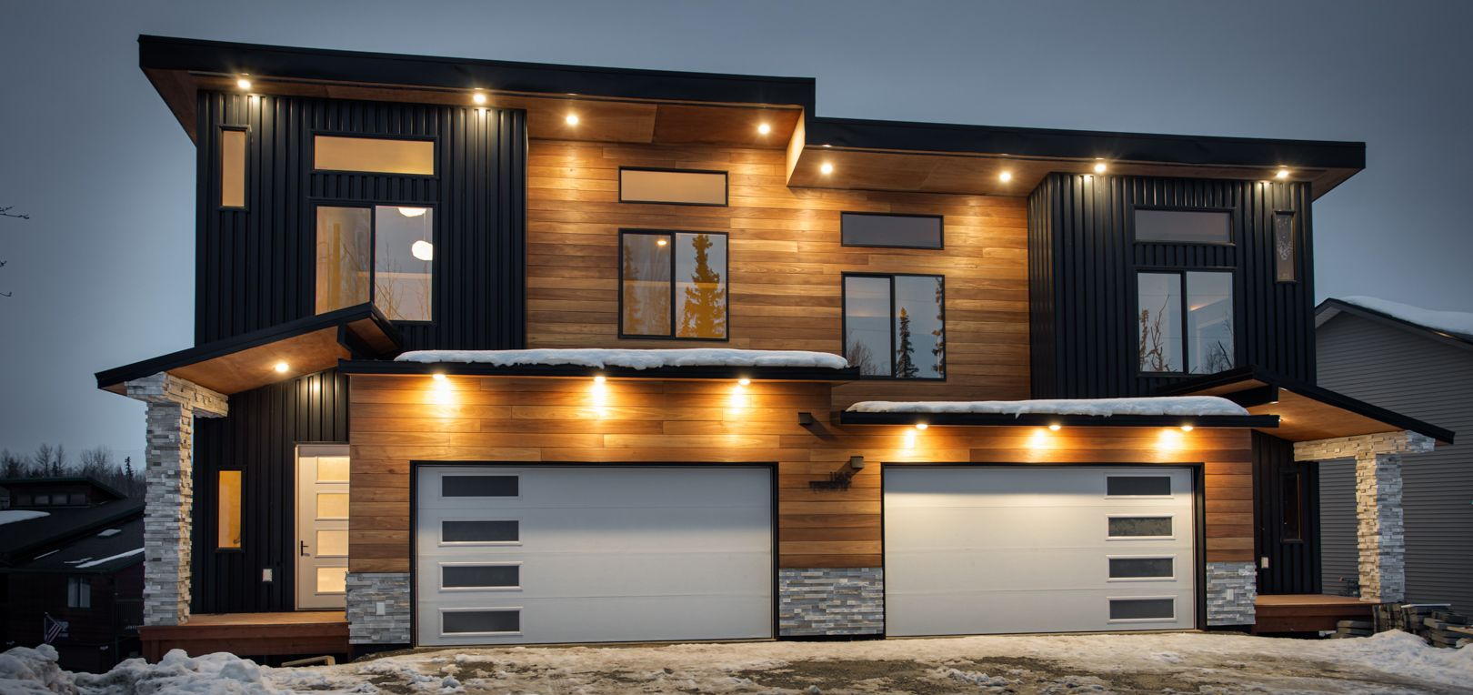 A modern two-story home with wood siding, black metal accents, and two garage doors at dusk, surrounded by light snow.