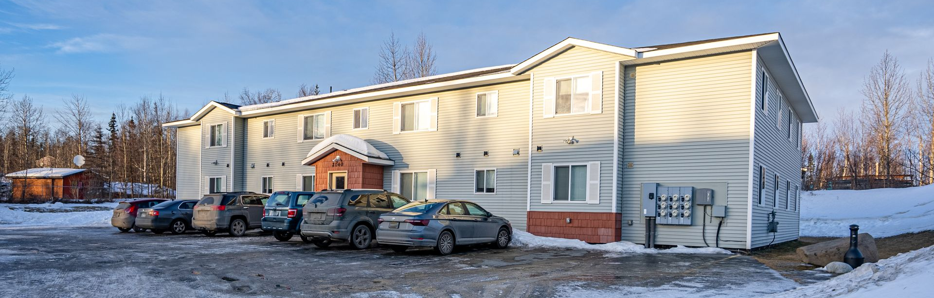 A light-colored two-story apartment building with a parking lot in front covered in snow under a clear blue sky.