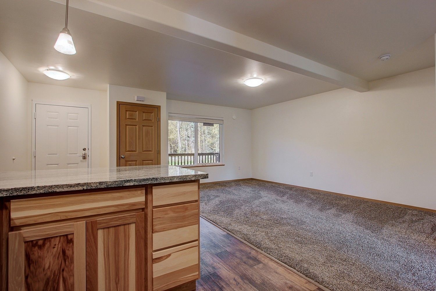 A kitchen counter with wood cabinets, transitioning to a living area with carpet and a window overlooking a deck.