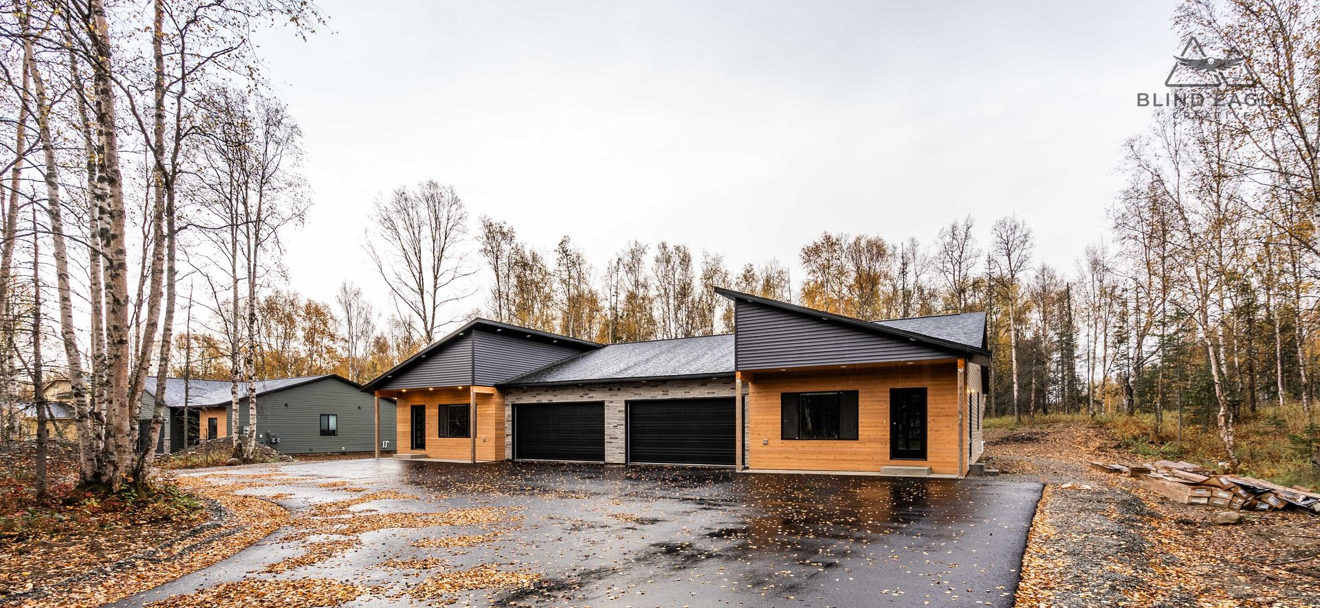 Modern wooden duplex with black slanted roofs, matching garages, and wet asphalt driveway surrounded by autumn trees.