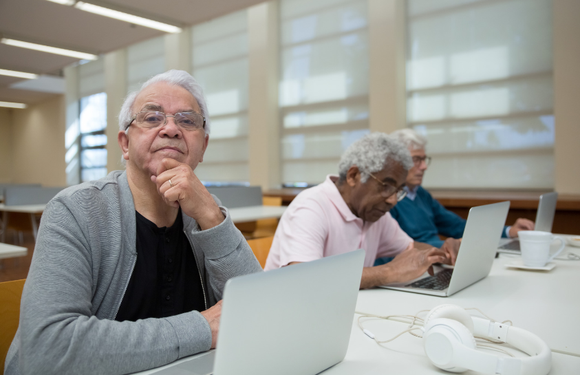A group of older men are sitting at a table using laptops.