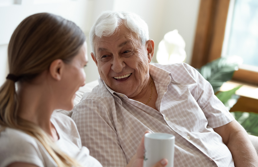 An elderly man is sitting on a couch talking to a young woman who is holding a cup of coffee.
