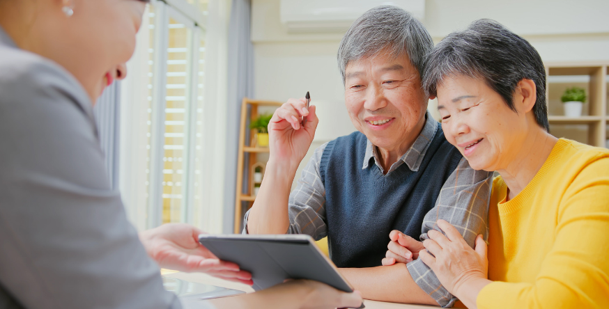 An elderly couple is sitting at a table looking at a tablet.
