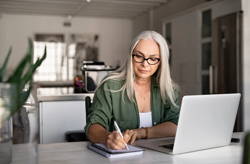 A woman is sitting at a table using a laptop computer and writing in a notebook.