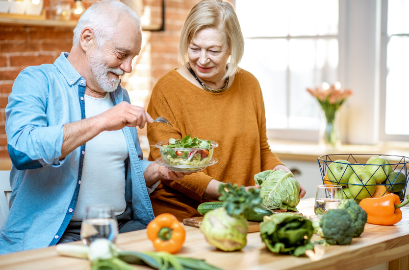 An elderly couple is preparing a salad in a kitchen.