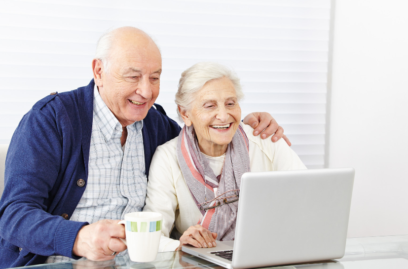 An elderly couple is sitting at a table looking at a laptop computer.