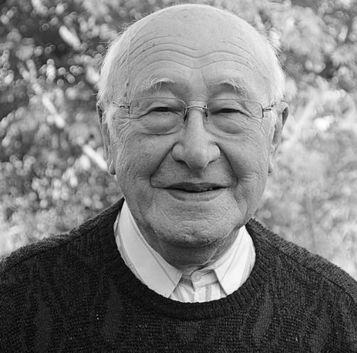 An elderly man wearing glasses and a sweater is smiling in a black and white photo.